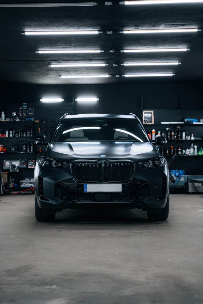 BMW SUV gris mat : Révélation d'un detailing auto parfait SUV BMW gris mat, plaque PL, garé dans un studio de detailing avec étagères de produits. Lumières néon reflétées sur la carrosserie.