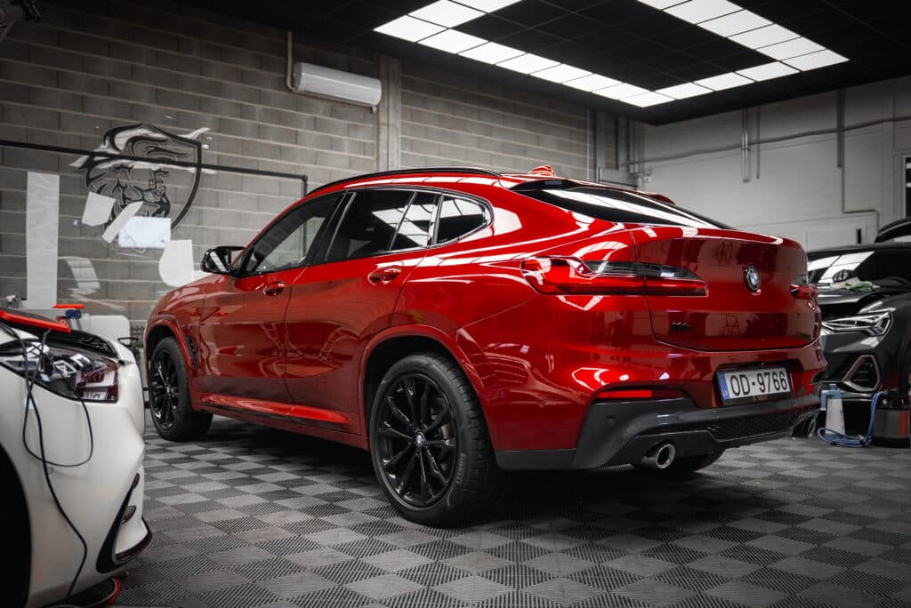 BMW X4 Rouge: Detailing Haut de Gamme en Atelier Pro BMW X4 rouge brillante en atelier de detailing, jantes noires, carrosserie reflétant l'environnement. Plancher à carreaux, mur en briques grises.