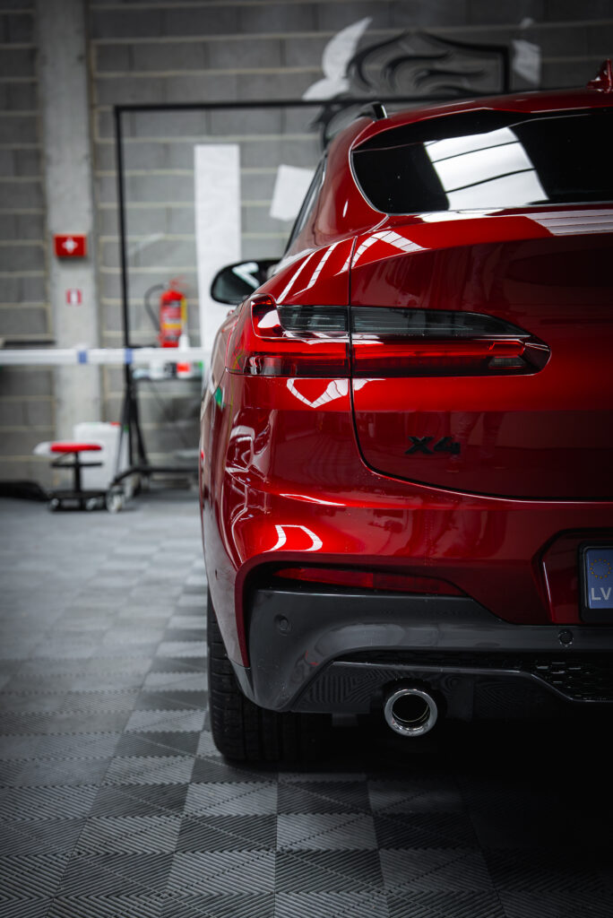 BMW X4 Rouge : Détailing de Luxe et Brillance Parfaite BMW X4 rouge rutilant, vue arrière. Badge 'X4' et plaque 'LV' visibles. Carrosserie brillante reflétant l'atelier moderne aux murs de parpaings.