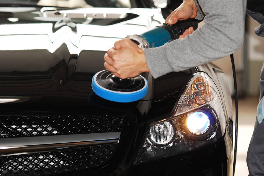 Polissage voiture noire : Rénovation et detailing auto expert Personne polissant le capot d'une voiture noire brillante avec une lustreuse électrique à pad bleu. Le phare de la voiture est allumé.